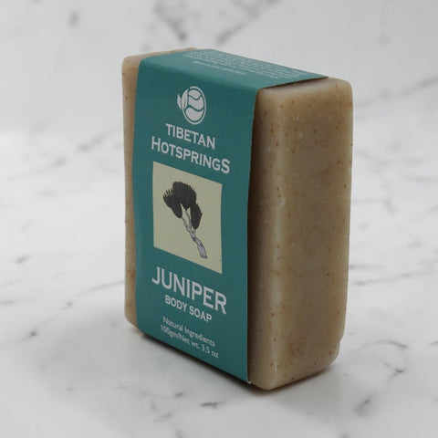 House of Nepal Tibetan Hotsprings Juniper Body Soap, eine feste, hellbraune Seife mit natürlichen Einschlüssen, umhüllt von einem türkisfarbenen Papierband mit Wacholderzweig-Illustration.