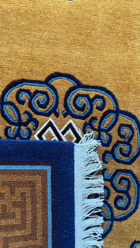 House of Nepal Teppich aus tibetischer Wolle in Ockergelb mit blau-weißen Endlosschleifen-Motiven und Fransen.